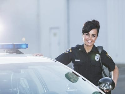 A policewoman standing next to her patrol car, smiling and looking at the camera with her hands on her hips. She is wearing the standard navy blue policeuniform.